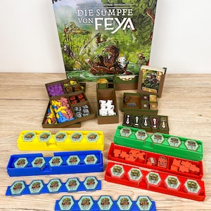May include: A board game titled Die Sümpfe von Feya with a large illustrated box, brown plastic component organisers, and colourful trays in yellow, blue, green, and red. The set includes various hexagonal tiles, small wooden tokens, and cards.