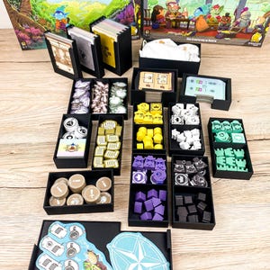 Könnte beinhalten: Ein Brettspiel namens "Navoria" mit Spielsteinen und Komponenten, die in schwarzen Tabletts angeordnet sind. Das Spiel enthält Karten, Spielsteine und Holzstücke in Farben wie Gelb, Lila und Grün. Die Spielbox ist im Hintergrund sichtbar, mit dem Titel auf Englisch und Deutsch.