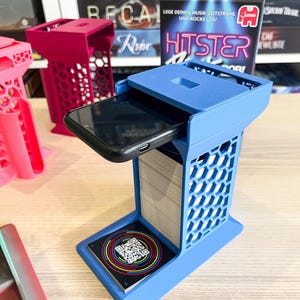 Pode incluir: Um suporte de telefone azul impresso em 3D com um design de favo de mel. Um smartphone preto repousa na bandeja superior, e uma pilha de cartões é visível no interior. Um código QR é impresso em um cartão na base. Outros suportes em rosa e magenta estão no fundo.