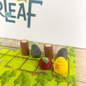 Puede incluir: Un juego de mesa con un diseño de campo verde, con piezas de tronco de madera, piedras grises, una manzana roja con una hoja verde y una colmena amarilla. El juego de mesa tiene la palabra "LEAF" en letras azules.