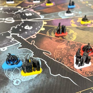 Puede incluir: Un juego de mesa con un tablero oscuro e ilustrado que presenta varias piezas de juego con forma de ciudad en negro, rojo, amarillo, azul y blanco. Las piezas del juego se colocan en el tablero, que tiene un diseño similar a un mapa con números y símbolos.