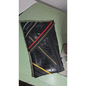 May include: A black clutch bag with a crocodile skin texture, featuring multiple diagonal zippers in black, red, and yellow. The bag has a rectangular shape with a fold-over design and metallic zipper teeth accents across the front surface.
