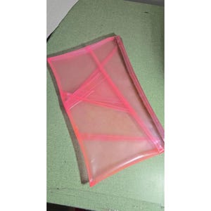 Puede incluir: Un estuche rectangular semitransparente de color rosa neón hecho de un material plástico flexible. El estuche presenta una serie de costuras estructurales diagonales y superpuestas que crean un patrón geométrico en su superficie.