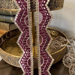 Op de afbeelding: Twee strengen van maroon en gouden trim met gespiegelde pailletten. De trim heeft een gegolfde rand en is perfect om een ​​toets van glans toe te voegen aan elk project.
