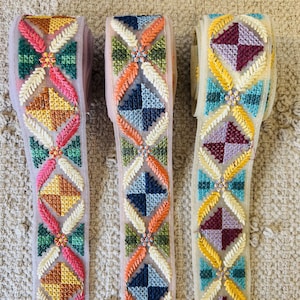 Op de afbeelding: Drie geborduurde lintversieringen met verschillende kleurcombinaties. De versieringen hebben een geometrisch patroon met een centraal bloemenmotief. De kleuren omvatten roze, oranje, geel, groen, blauw en rood.