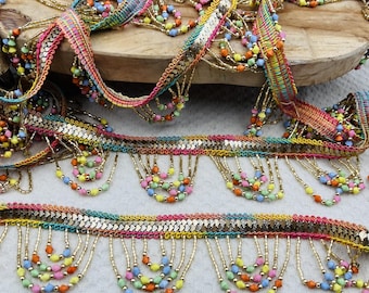 Könnte beinhalten: Ein dekoratives Band mit einem geflochtenen goldfarbenen Metallstreifen und mehrfarbigen gewebten Kanten in Pink, Gelb und Blau. Das Band ist mit hängenden Fransen aus goldfarbenen Perlen und kleinen, bunten facettierten Perlen in Pastelltönen verziert.