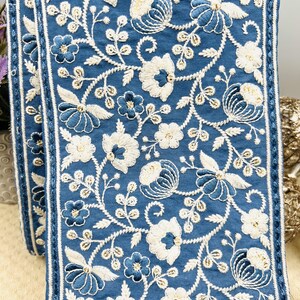 K&ouml;nnte beinhalten: Blaues, besticktes Stoffband mit wei&szlig;en und blauen Blumenmustern. Der Besatz weist detaillierte Stickereien verschiedener Blumen und Bl&auml;tter mit strukturiertem Aussehen auf. Die Kanten sind mit einem d&uuml;nnen wei&szlig;en Streifen eingefasst.
