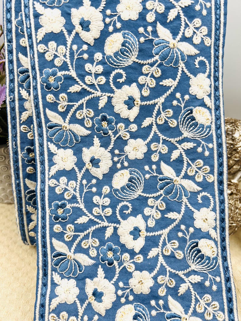 K&ouml;nnte beinhalten: Blaues, besticktes Stoffband mit wei&szlig;en und blauen Blumenmustern. Der Besatz weist detaillierte Stickereien verschiedener Blumen und Bl&auml;tter mit strukturiertem Aussehen auf. Die Kanten sind mit einem d&uuml;nnen wei&szlig;en Streifen eingefasst.