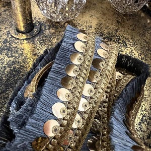May include: Close-up of decorative trim with gold sequins and fringe. The trim is a combination of gold and grey-blue, with a textured appearance. The sequins are circular and add a touch of sparkle. The fringe is soft and flowing.
