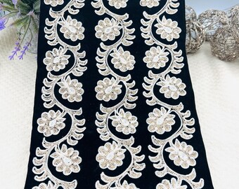 Könnte beinhalten: Schwarzer Stoff mit drei vertikalen Reihen weißer und goldener Blumenstickerei. Das florale Design zeigt detaillierte Blumen und Blätter. Der Stoff ist gefaltet und zeigt die Textur und das Muster. Diese dekorative Borte eignet sich für Bastelprojekte.