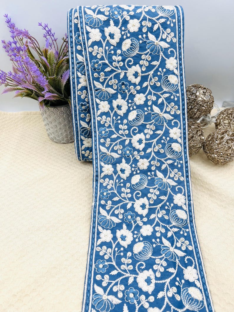 K&ouml;nnte beinhalten: Eine Rolle blaues Stoffband mit wei&szlig;er Blumenstickerei. Das Band weist ein sich wiederholendes Muster aus Blumen und Bl&auml;ttern auf. Die Kanten sind mit einem d&uuml;nnen wei&szlig;en Rand versehen. Das Band wird wahrscheinlich f&uuml;r Bastel- oder Dekorationszwecke verwendet.