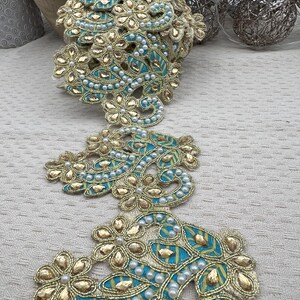 Könnte beinhalten: Dekorative Zierleiste mit einem floralen Design. Die Zierleiste hat eine goldfarbene metallische Kontur mit türkisfarbenen und gelben Akzenten. Kleine weiße Perlen sind über das Design verstreut. Diese Zierleiste wird wahrscheinlich zum Verschönern von Kleidung oder Bastelprojekten verwendet.