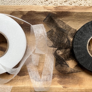 Op de afbeelding: Twee rollen stoffen tape, een witte en een zwarte, worden getoond op een houten oppervlak. De witte tape is een smeltbaar bindweefsel, en de zwarte tape is een naai-interfacing.
