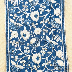 K&ouml;nnte beinhalten: Nahaufnahme eines Textils mit einer zentralen blauen Fl&auml;che, die mit wei&szlig;en und blauen Blumenstickereien verziert ist. Die Stickerei umfasst verschiedene Blumenmotive und Blattranken. Die blaue Fl&auml;che wird von einem wei&szlig;-blauen, gen&auml;hten Rand vor einem beigen Hintergrund umrahmt.