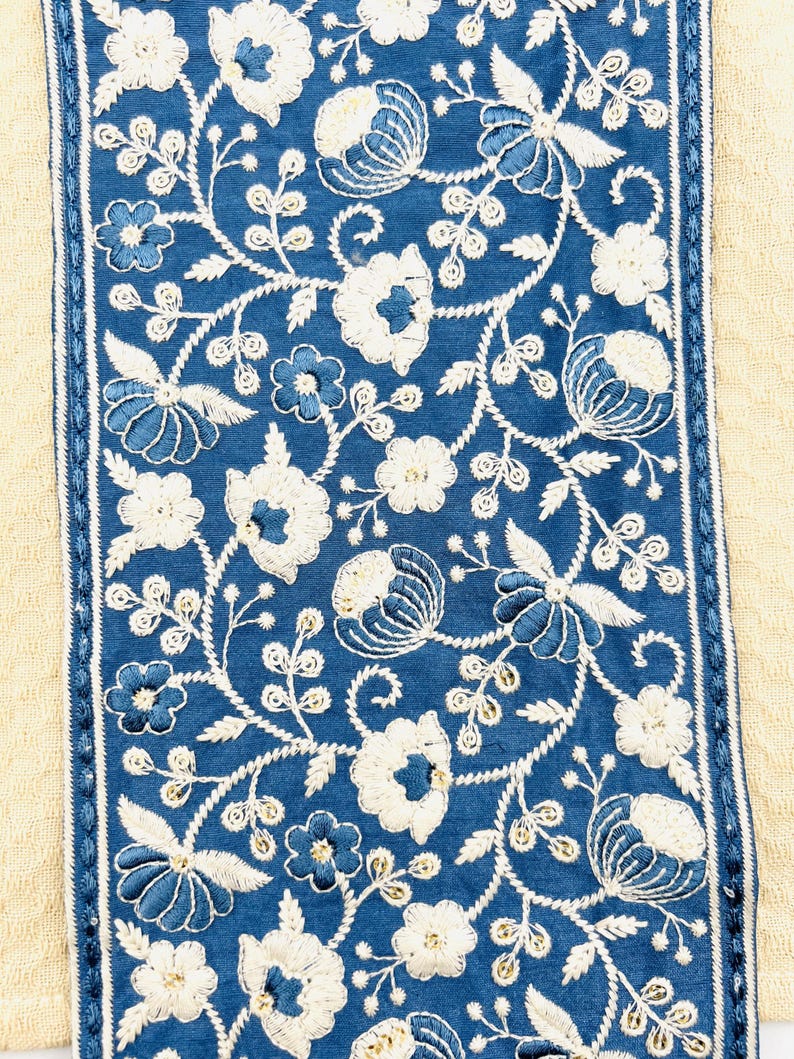 K&ouml;nnte beinhalten: Nahaufnahme eines Textils mit einer zentralen blauen Fl&auml;che, die mit wei&szlig;en und blauen Blumenstickereien verziert ist. Die Stickerei umfasst verschiedene Blumenmotive und Blattranken. Die blaue Fl&auml;che wird von einem wei&szlig;-blauen, gen&auml;hten Rand vor einem beigen Hintergrund umrahmt.