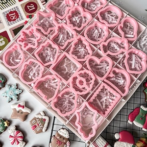 K&ouml;nnte beinhalten: Ein Set rosa Weihnachtspl&auml;tzchenausstecher in verschiedenen Formen, darunter Herzen und Sterne, angeordnet in einer unterteilten Box. Die Box ist mit rosa Papier gef&uuml;llt. Dekorierte Lebkuchenkekse sind um die Box herum verstreut.