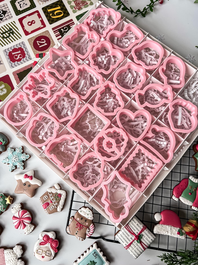 K&ouml;nnte beinhalten: Ein Set rosa Weihnachtspl&auml;tzchenausstecher in verschiedenen Formen, darunter Herzen und Sterne, angeordnet in einer unterteilten Box. Die Box ist mit rosa Papier gef&uuml;llt. Dekorierte Lebkuchenkekse sind um die Box herum verstreut.
