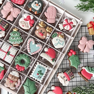 K&ouml;nnte beinhalten: Eine Box mit dekorierten Weihnachtspl&auml;tzchen, darunter ein Strumpf, ein Pinguin, ein F&auml;ustling, eine Schleife und der Weihnachtsmann. Die Kekse sind in verschiedenen Farben gehalten, darunter Rot, Rosa, Gr&uuml;n und Wei&szlig;. Einige Kekse haben den Text "GOD JUL" und "30&cent;".