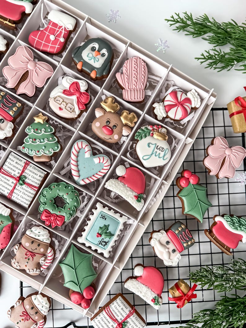 K&ouml;nnte beinhalten: Eine Box mit dekorierten Weihnachtspl&auml;tzchen, darunter ein Strumpf, ein Pinguin, ein F&auml;ustling, eine Schleife und der Weihnachtsmann. Die Kekse sind in verschiedenen Farben gehalten, darunter Rot, Rosa, Gr&uuml;n und Wei&szlig;. Einige Kekse haben den Text "GOD JUL" und "30&cent;".