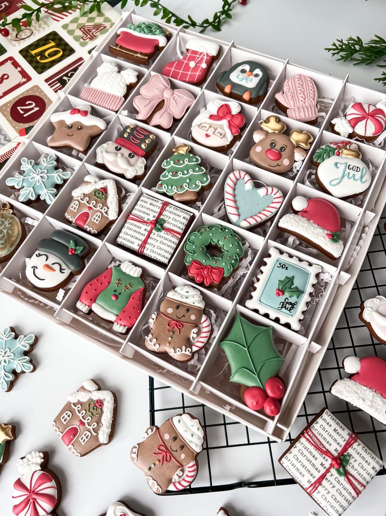 K&ouml;nnte beinhalten: Eine Schachtel mit dekorierten Weihnachtspl&auml;tzchen, darunter Lebkuchenm&auml;nner, Weihnachtsm&uuml;tzen und Schneeflocken. Die Kekse sind in verschiedenen Formen und Farben gehalten und weisen aufwendige Verzierungen auf. Einige Kekse tragen den Text "GOD JUL".