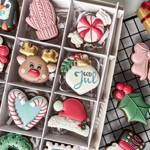 K&ouml;nnte beinhalten: Eine Schachtel mit dekorierten Weihnachtspl&auml;tzchen, darunter ein Pinguin, ein rosa F&auml;ustling, eine Zuckerstange, ein Rentier und eine Weihnachtsm&uuml;tze. Weitere Kekse sind ein Herz, ein Kranz und eine Briefmarke. Die Kekse befinden sich in einer wei&szlig;en Box.