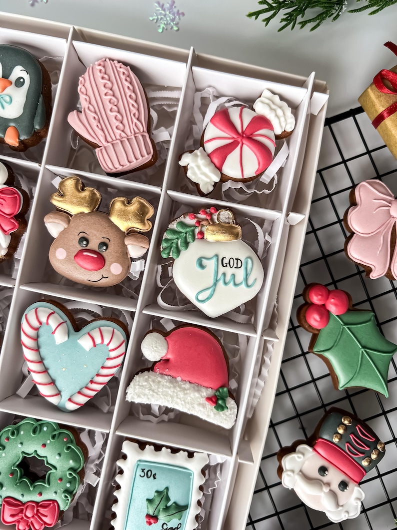 K&ouml;nnte beinhalten: Eine Schachtel mit dekorierten Weihnachtspl&auml;tzchen, darunter ein Pinguin, ein rosa F&auml;ustling, eine Zuckerstange, ein Rentier und eine Weihnachtsm&uuml;tze. Weitere Kekse sind ein Herz, ein Kranz und eine Briefmarke. Die Kekse befinden sich in einer wei&szlig;en Box.