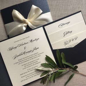 May include: A black and ivory wedding invitation with a white satin ribbon bow. The invitation includes the names Tiffany Andres Mendoza and Joshua Camejo Stogpos, the date and time of the wedding ceremony, and the location of the reception. The text "Entourage", "Gifting", and "RSVP" is also included on the invitation.