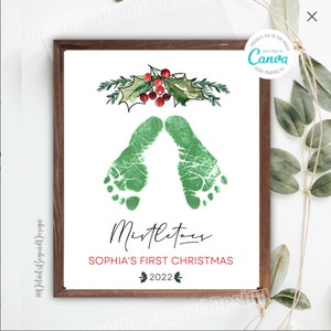 May include: A framed print featuring green baby footprints, holly, and the text "Mistletoes, Sophia's First Christmas 2022." The print is in a brown wooden frame. A Canva logo is in the upper right corner.