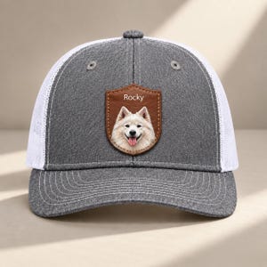 Op de afbeelding: Een grijs-witte truckerpet met een bruine, leerachtige schildvormige patch op de voorkant. De patch toont de naam Rocky boven een gedetailleerde illustratie van een witte, pluizige hond met de tong uit de bek op het voorpaneel van de pet.