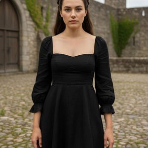 May include: A black dress with a square neckline and long sleeves. The dress has a fitted bodice and a flared skirt. The sleeves have a ruffled detail at the wrist. The dress is made of a solid black fabric.