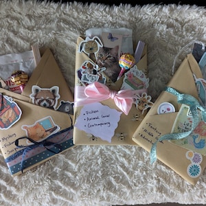 Könnte beinhalten: Drei braune Pakete aus Packpapier, die mit Schleife und Aufklebern verziert sind, darunter Tieraufkleber, ein Meerjungfrauenaufkleber und ein Aufkleber mit dem Text "Let's be Mermaids". Die Pakete sind mit handschriftlichen Notizen versehen, die das Genre der Bücher im Inneren angeben: "Mystery, Fiction, Suspense", "Fiction, Tierliebhaber, Zeitgenössisch" und "Fantasy, Middle Grade, Abenteuer".