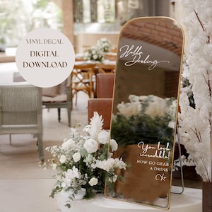 Puede incluir: Un espejo con marco dorado con una calcomanía de vinilo blanco que dice "Hello Darling" y "You look incredible! Now go grab a drink XO". El espejo está sobre una superficie blanca con flores blancas delante.