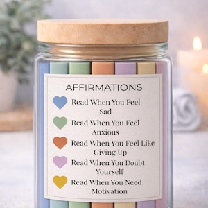May include: A glass jar with a wooden lid, filled with colorful paper strips. The jar has a label that reads "AFFIRMATIONS" and lists affirmations for various emotional states. The jar is a self-care or wellness product.