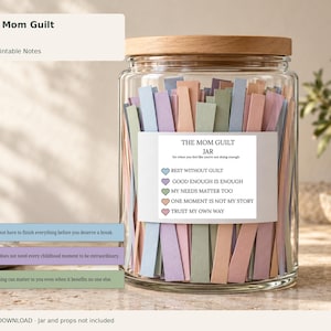 Puede incluir: Un frasco de vidrio transparente con tapa de madera lleno de tiras de papel multicolor. Una etiqueta blanca en el frente dice THE MOM GUILT JAR con cinco iconos de corazón y frases que incluyen REST WITHOUT GUILT y TRUST MY OWN WAY.