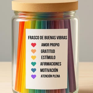 Op de afbeelding: Een glazen pot met een houten deksel, gevuld met kleurrijke papieren stroken. Een wit etiket op de pot leest "FRASCO DE BUENAS VIBRAS" met affirmaties. De papieren stroken zijn in regenboogkleuren gerangschikt.