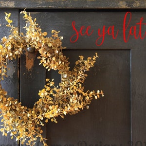 May include: A dried, golden-colored wreath hangs on a dark wooden door with the text "see ya later" written in red above it. The wreath is made of small, delicate leaves and branches.