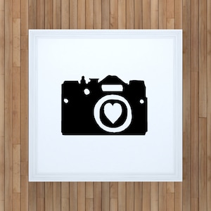 May include: Black and white silhouette of a camera with a heart in the lens. The camera is framed in a white frame.