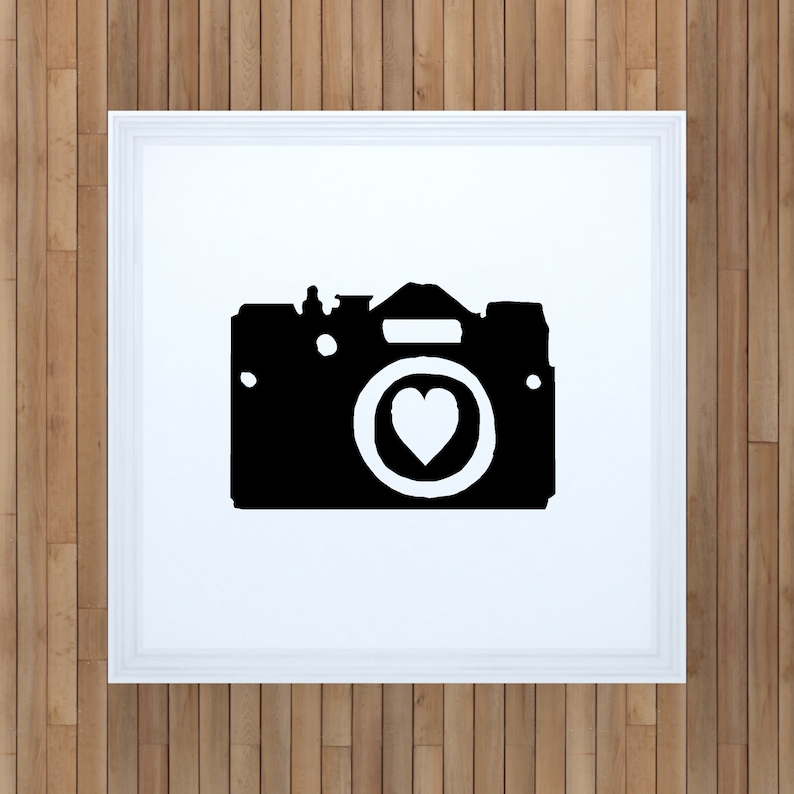 May include: Black and white silhouette of a camera with a heart in the lens. The camera is framed in a white frame.