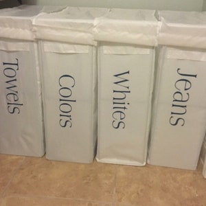 May include: Four white laundry hampers with blue lettering. The hampers are labeled "Towels", "Colors", "Whites", and "Jeans".