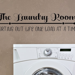 Op de afbeelding: Een witte wasmachine met een zwarte tekst sticker die "The Laundry Room Sorting Out Life One Load At A Time" zegt boven de machine.