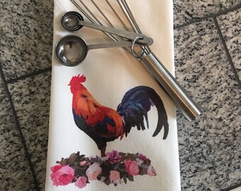 May include: White kitchen towel with a colorful rooster illustration and pink flowers. A silver whisk and measuring spoons are on top of the towel.