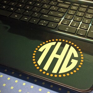 Op de afbeelding: Een zwarte laptop met een gele en witte monogram sticker met de tekst "THE" in een cirkel.