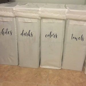 May include: Four white laundry hampers with black labels. The labels read "whites", "darks", "colors", and "towels".