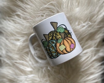 Può includere: Tazza in ceramica bianca con un'illustrazione disegnata a mano di tre zucche. Le zucche sono in tonalità di verde, marrone e arancione. Le zucche hanno un design a patchwork con quadrati di diversi colori.