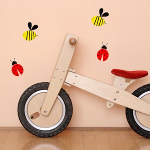 Puede incluir: Una bicicleta de equilibrio de madera con neumáticos negros y un asiento rojo. La bicicleta está delante de una pared rosa claro con pegatinas de abejas negras y amarillas y mariquitas rojas.