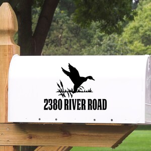 May include: White mailbox with a black silhouette of a duck landing near reeds. The text "2380 RIVER ROAD" is printed in black below the duck graphic. The mailbox is mounted on a wooden post.