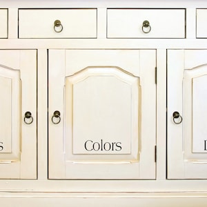 Op de afbeelding: Witte kast met drie deuren met de labels "Witten", "Kleuren" en "Donkers". Elke deur heeft een decoratief gebogen paneel en een metalen knop.
