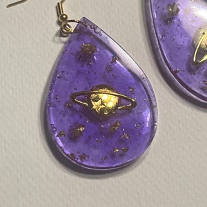 Peut inclure: Boucles d'oreilles en forme de goutte d'eau violettes avec un motif de planète doré. Les boucles d'oreilles sont remplies de paillettes dorées et ont des crochets d'oreilles dorés. Un accessoire unique.