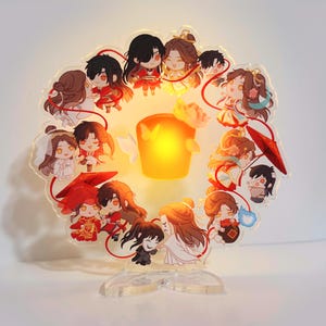 May include: An acrylic lamp with a circular design, featuring cartoon characters in various outfits. The lamp has a warm, yellow light in the centre and a clear base. The characters are in red, white, and gold.