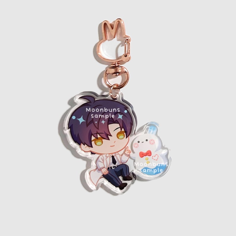 May include: An acrylic keychain featuring a chibi-style character with dark purple hair and yellow eyes, wearing a white coat and dark pants. Next to the character is a small white ghost-like figure with a red bow tie. It has a rose gold rabbit-shaped clasp.